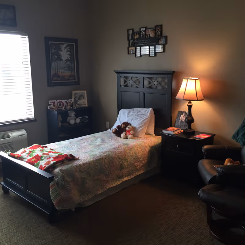 A cozy bedroom with a single bed covered in a colorful quilt and two stuffed animals on the pillow. Next to the bed is a nightstand with a lit table lamp, books, and a framed photo. There is a dark wooden armchair to the right and a small bookshelf with decorative items and photos near a window with blinds. The walls are painted beige and decorated with framed pictures.
