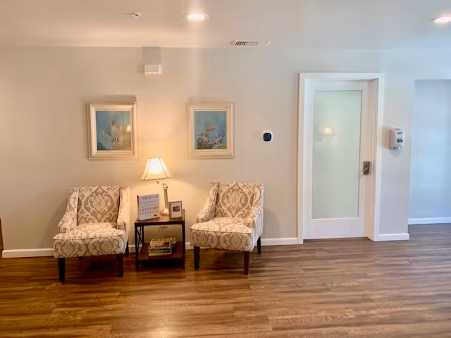 A cozy interior seating area with two patterned armchairs separated by a small table holding a lamp, books, and a framed photo. Two framed paintings hang on the beige wall above the chairs. To the right, there is a white door with frosted glass and a wall-mounted hand sanitizer dispenser. The floor is wooden.
