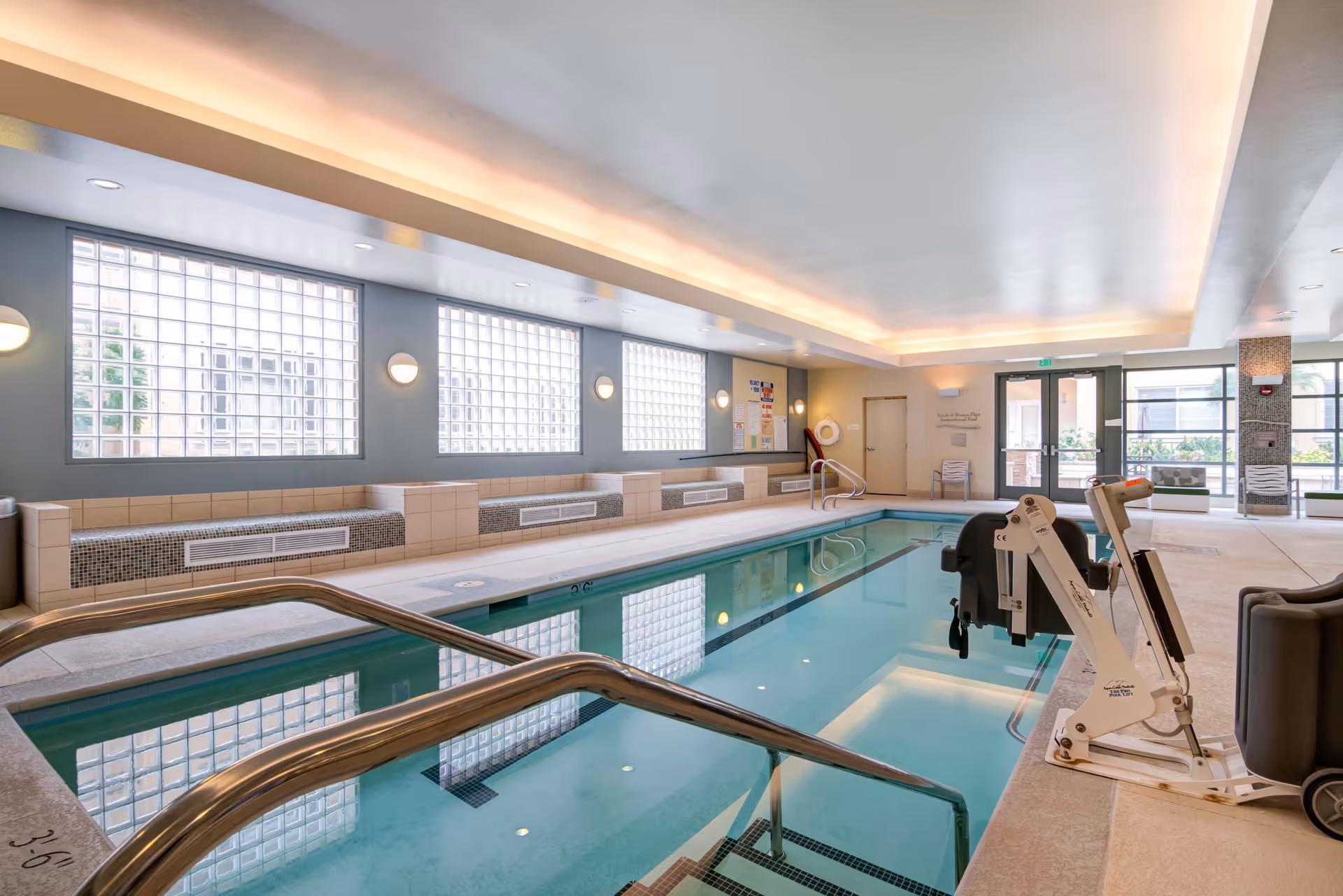 Indoor swimming pool area with clear water, metal handrails, and a pool lift chair for accessibility. The room has large glass block windows allowing natural light, tiled walls with built-in bench seating, and a ceiling with recessed lighting. There is a door and glass exit doors leading outside in the background.