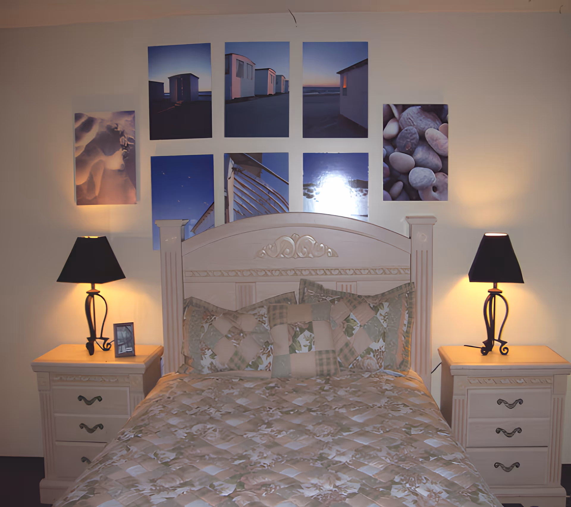 A bedroom with a wooden bed featuring a carved headboard and a floral patchwork quilt. There are two matching nightstands on either side of the bed, each with a black lampshade table lamp. Above the bed, there are eight framed pictures arranged in two rows, depicting various scenes including beach huts, pebbles, and abstract images.