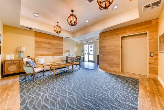 A senior living facility lobby with sofas, chairs and a coffee table under pendant lights, patterned carpet and an elevator near the entrance.