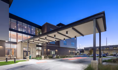 Exterior view of a modern senior living facility entrance at dusk with a covered drop-off area supported by columns, large windows, and a well-lit driveway.