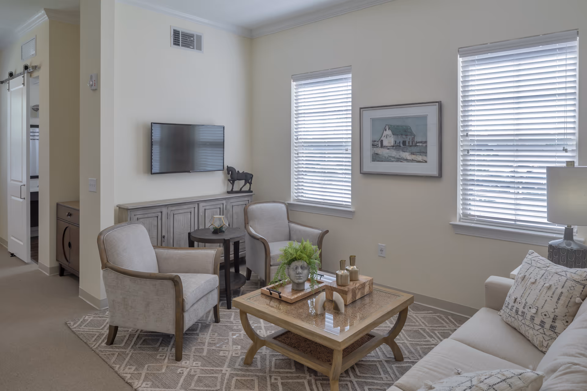 A cozy living room with two beige armchairs, a beige sofa with patterned pillows, a wooden coffee table with decorative items including a plant and small sculptures, a TV mounted on the wall above a wooden cabinet, two windows with white blinds, a framed picture on the wall, and a lamp on a side table.