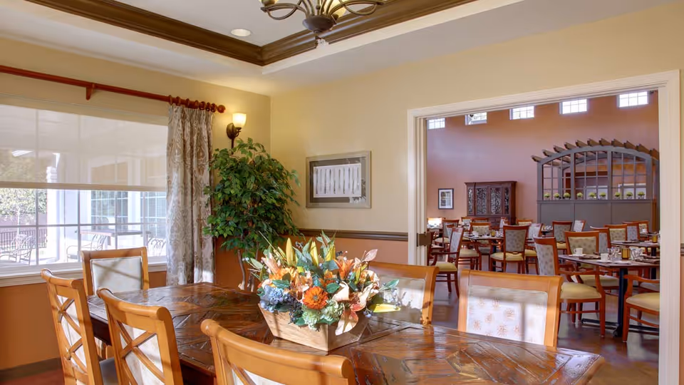 A well-lit dining area with a wooden table and chairs in the foreground, decorated with a floral centerpiece. In the background, another dining room is visible with multiple tables and chairs arranged neatly, warm-colored walls, and large windows letting in natural light.