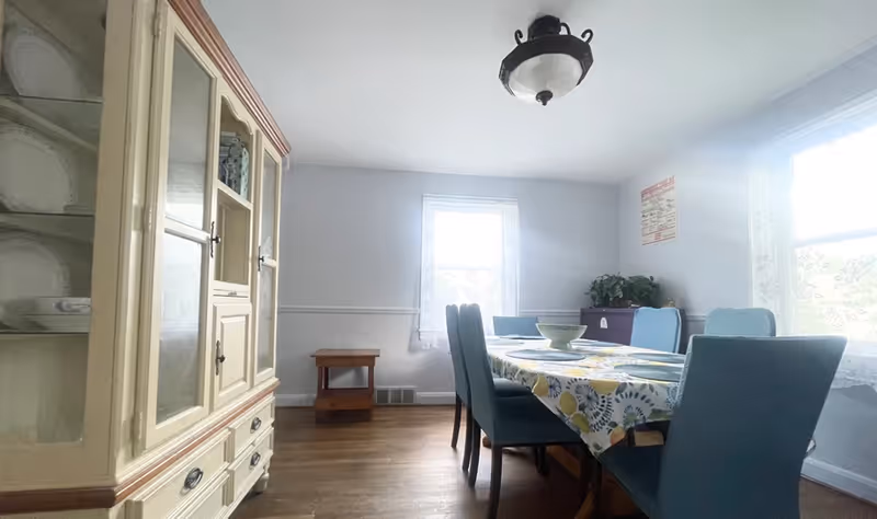 A dining room with a wooden floor, a table covered with a colorful tablecloth, six blue upholstered chairs around the table, a cream-colored cabinet with glass doors on the left, two windows with lace curtains letting in natural light, and a small wooden side table against the far wall.