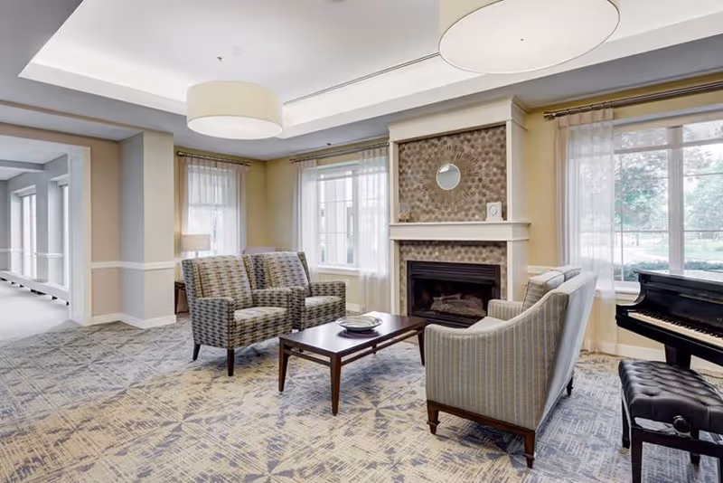 A cozy living room area in Buckingham's Choice featuring two patterned armchairs and a striped sofa arranged around a wooden coffee table. The room has a fireplace with a decorative tile surround and a small round mirror above it. Large windows with sheer curtains allow natural light to fill the space. A black piano with a cushioned bench is positioned near one window. The room has a patterned carpet and modern ceiling lights.