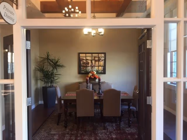 View through glass double doors into a private dining room with a rectangular wooden table surrounded by six upholstered chairs. A floral centerpiece is on the table, and a large potted plant is in the corner. A framed mirror hangs on the beige wall, reflecting a chandelier above the table.