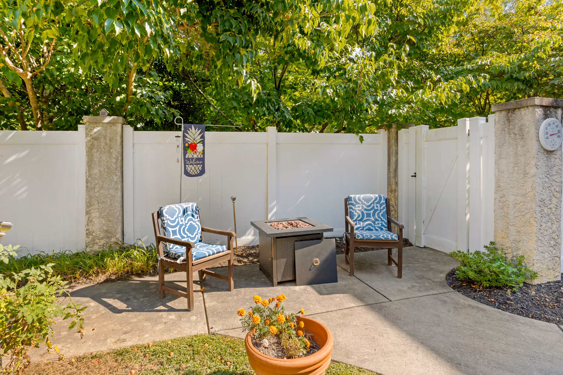 Outdoor patio area with two wooden chairs featuring blue and white patterned cushions, a square fire pit between them, a white fence in the background, green trees overhead, a welcome flag hanging on the fence, and a potted plant with yellow flowers in the foreground.