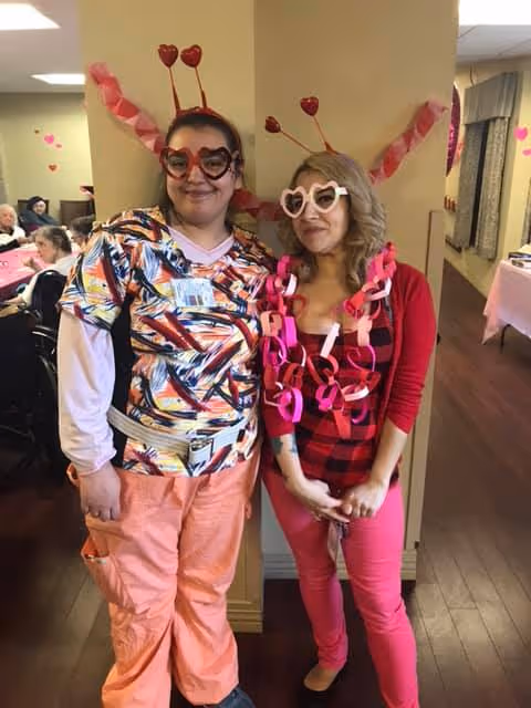 Two women standing indoors in a decorated room. Both are wearing heart-shaped glasses and headbands with heart antennae. One woman is dressed in colorful scrubs and peach pants, while the other wears a red plaid shirt, pink pants, and a red cardigan with paper chain decorations around her neck. In the background, several elderly people are seated at tables, suggesting a social or care facility setting.