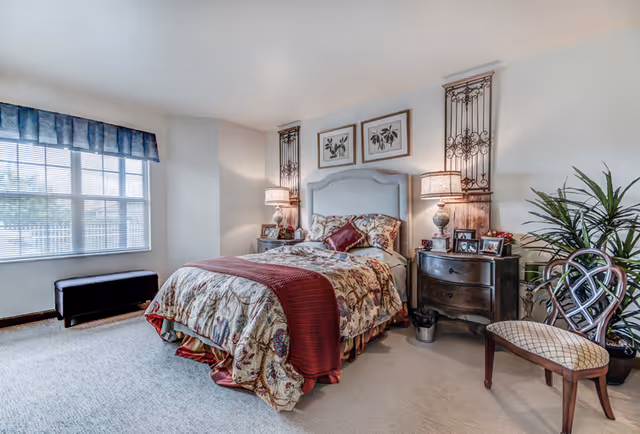 A well-appointed bedroom with a double bed dressed in patterned bedding, matching nightstands and lamps, a window bench, a chair, and a potted plant.