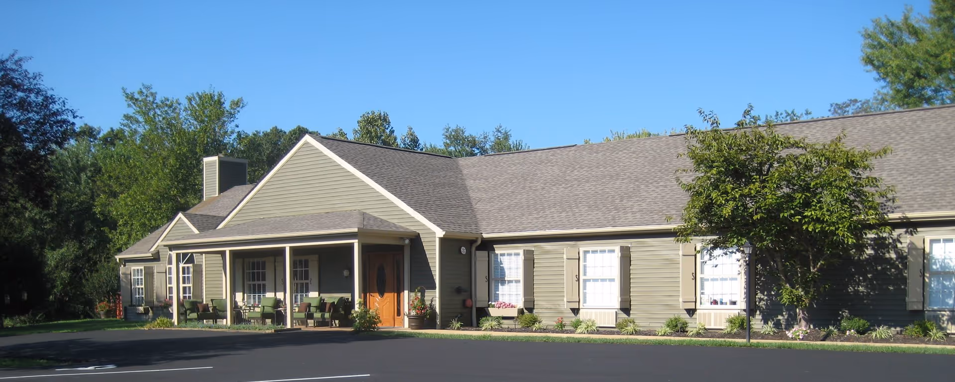 Exterior view of a single-story assisted living facility building with a covered porch, several chairs, and a well-maintained garden area. The building has green siding, white trim, and multiple windows with shutters. There is a paved parking area in front and trees surrounding the property.