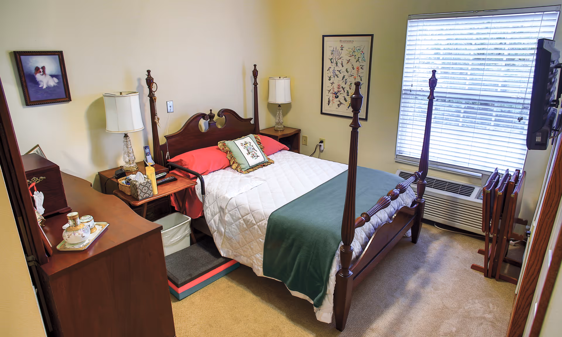 Neatly furnished bedroom with a four-poster bed, nightstands, dresser, window with blinds, and wall art.