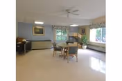 A spacious room with light-colored flooring and walls painted in a soft blue and beige. There is a round table with four chairs in the center, a ceiling fan above, and large windows with floral valances letting in natural light. The room also features framed artwork on the walls and a radiator beneath the windows.