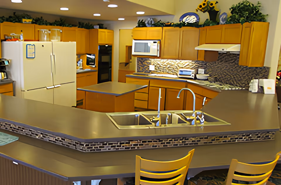 A spacious kitchen area with wooden cabinets, a double sink, a white refrigerator, a microwave, and an oven. The kitchen features a large countertop with a raised bar area and decorative plants and items on top of the cabinets.