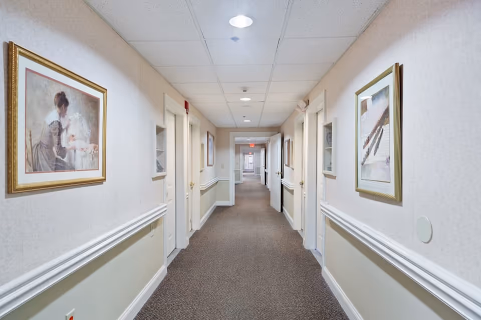 A long, well-lit hallway in a senior living facility with beige walls, white trim, and carpeted floor. Several closed white doors line both sides of the corridor, and framed artwork hangs on the walls. The ceiling has recessed lighting and a drop ceiling design.