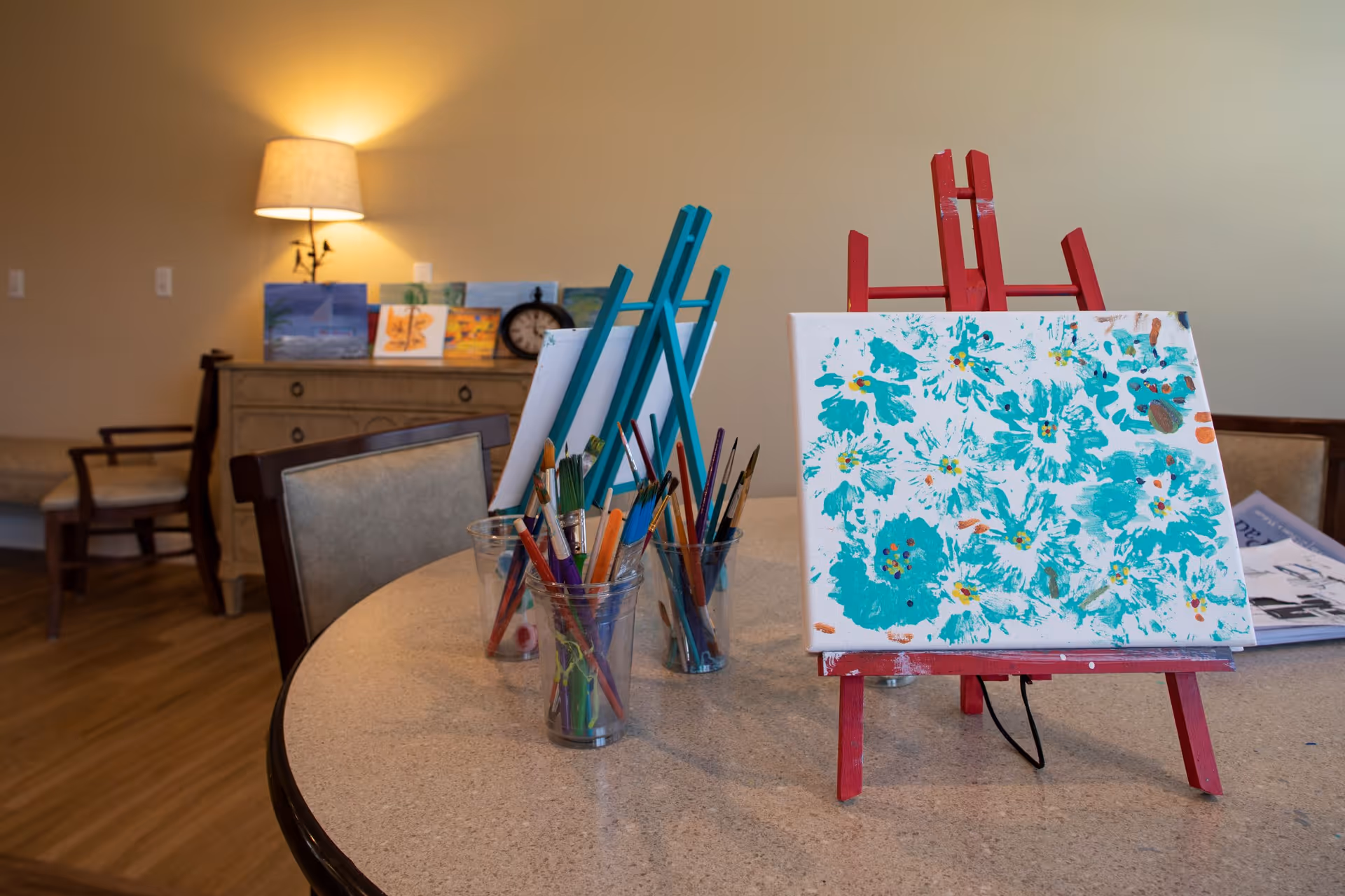 A round table with two small easels holding canvases, one with a painting of blue flowers. Several clear plastic cups filled with paintbrushes are on the table. In the background, there is a wooden sideboard with small paintings and a clock, a lit table lamp, and a chair with a cushion.