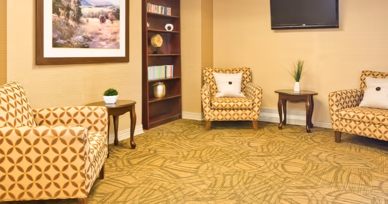 A cozy sitting area with three patterned armchairs arranged around two small wooden side tables. A bookshelf with books and decorative items is in the corner, and a flat-screen TV is mounted on the wall above one of the tables. A framed landscape painting hangs on the wall above one armchair. The room has warm beige walls and a patterned carpet.