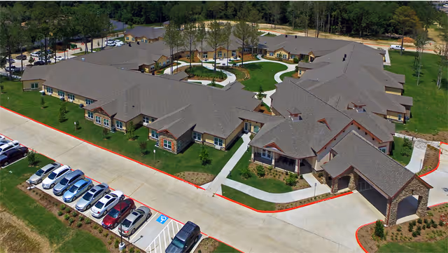 Aerial view of a large senior living community building with multiple connected sections, surrounded by green lawns and trees. There is a parking lot with several cars parked along a driveway in front of the building. Sidewalks and landscaped areas are visible around the facility.