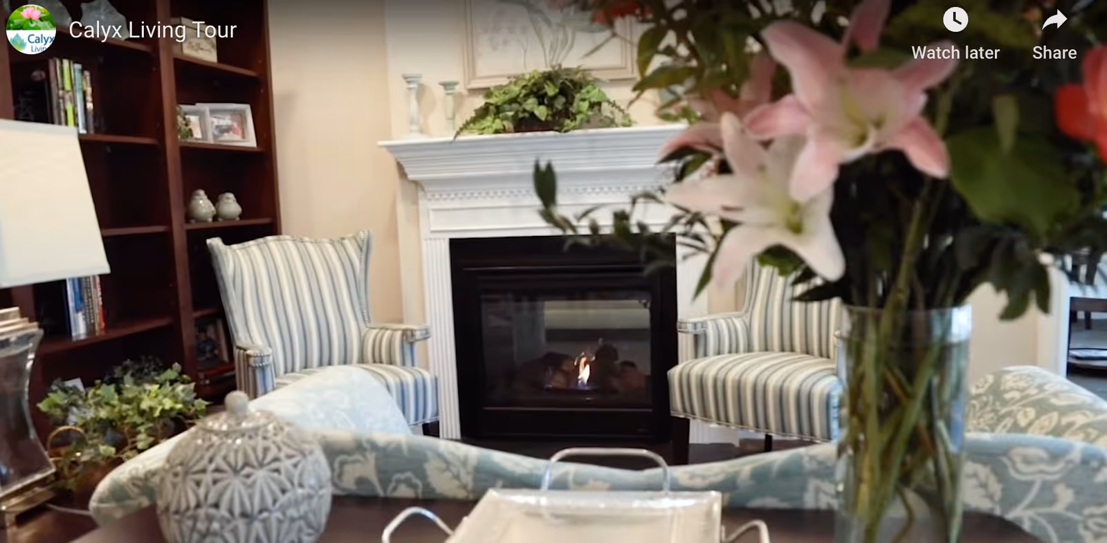 A cozy living room with a white fireplace in the center, two striped armchairs on either side, a wooden bookshelf filled with books and decorative items on the left, and a vase of pink and white flowers in the foreground.