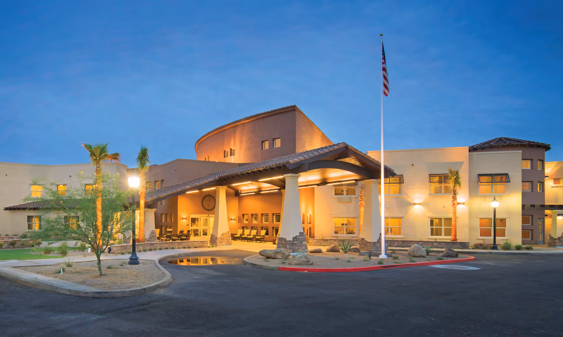 The lit front entrance of an assisted living building at dusk with a covered porte-cochere, flagpole, palm trees, and driveway.
