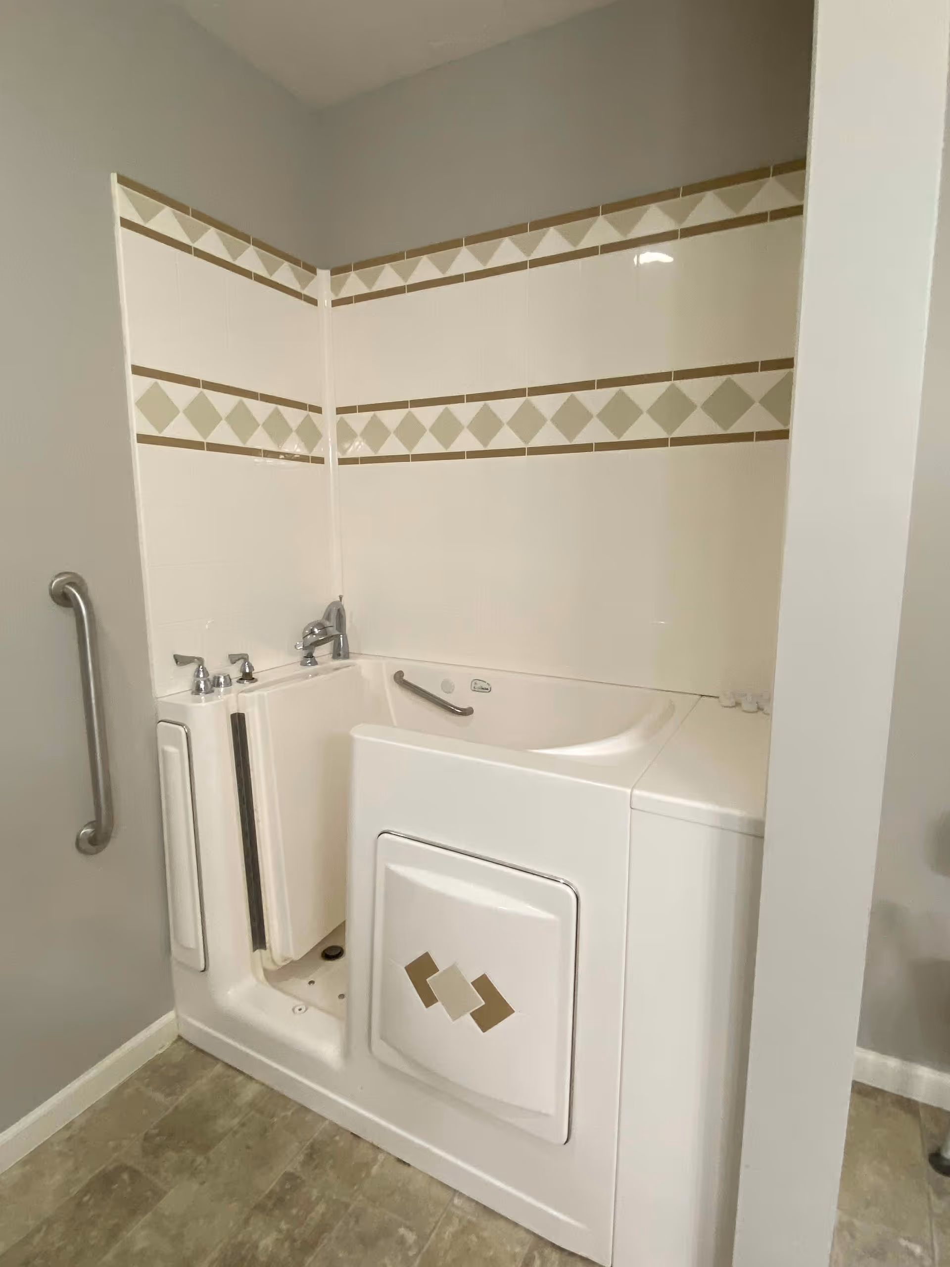 A walk-in bathtub with a door for easy access, equipped with a faucet and a safety grab bar. The surrounding walls are tiled with a decorative pattern of beige and light green diamonds and triangles. The floor has a stone-like tile pattern, and there is a metal grab bar mounted on the adjacent wall.