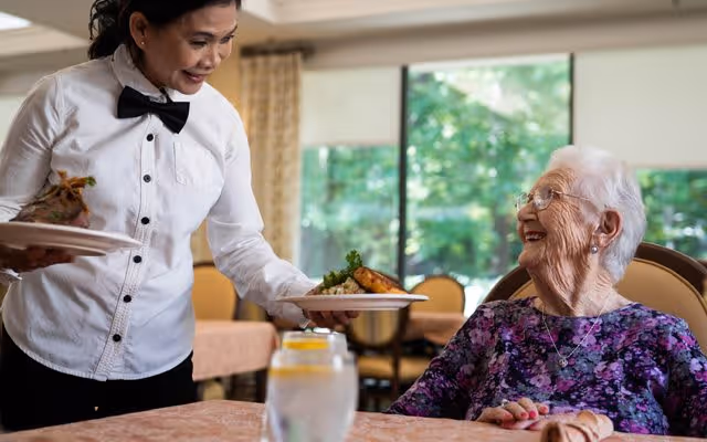 A smiling elderly woman with white hair and glasses sitting at a dining table in a senior living facility, looking up at a female server dressed in a white shirt and black bow tie who is serving a plate of food. The background shows large windows with greenery outside and other dining tables and chairs.
