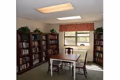 A small room with a rectangular wooden table and four wooden chairs. The room has three tall bookshelves filled with books and decorated with potted plants on top. There is a window with a red valance letting in natural light.