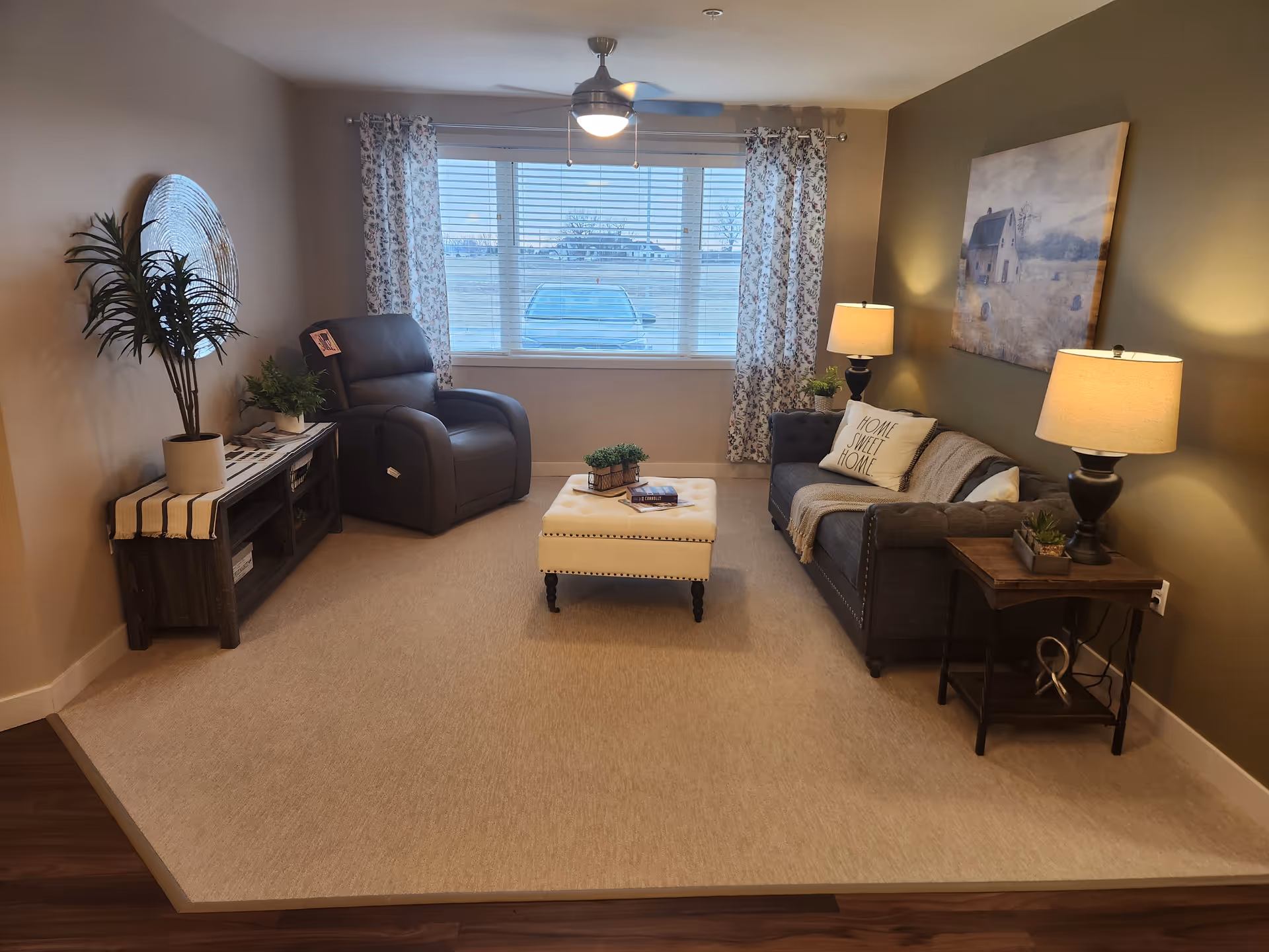 Bright living room with a sofa, recliner, ottoman, side tables and lamps arranged around a large window.