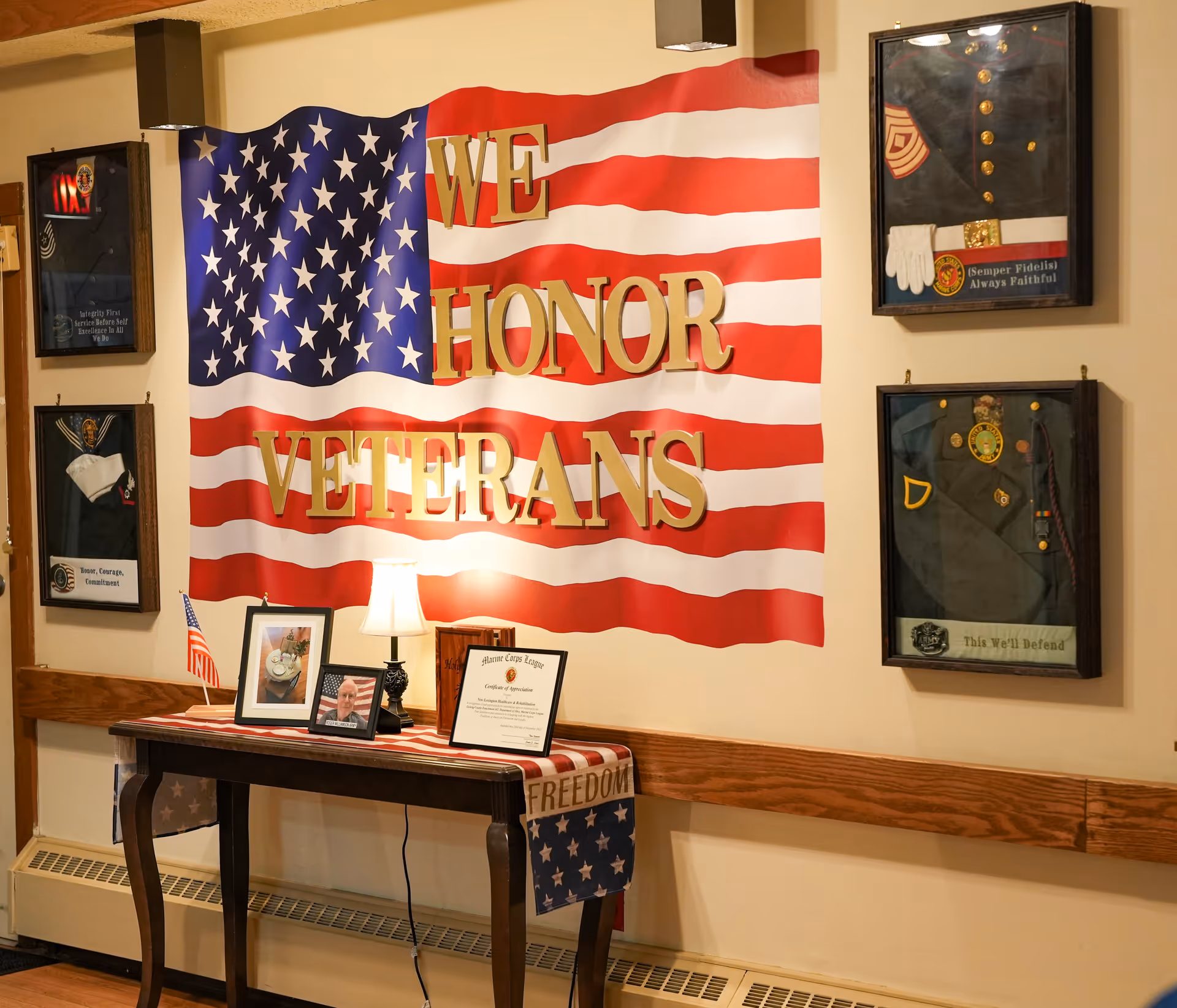 A wall display honoring veterans with a large American flag and gold letters spelling 'WE HONOR VETERANS'. Below the flag is a table with a small American flag, framed photos, a lamp, and a certificate. On the wall around the flag are four framed military uniforms and memorabilia.