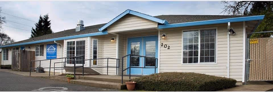 Exterior view of a single-story building with beige siding and blue trim, featuring a ramp and steps leading to a blue double door entrance. The building has multiple windows and the number 202 displayed near the door. There is a small bush and potted plants near the entrance, and a sign on the building reads 'Blue Haven Memory Care - Independence'.