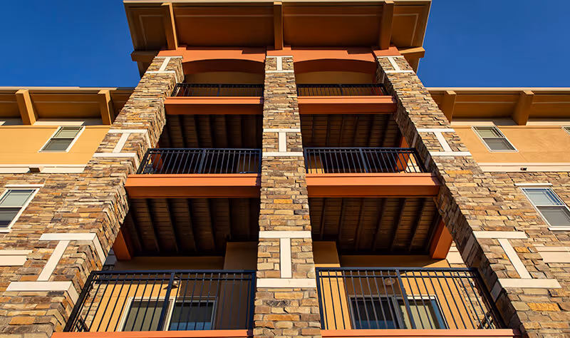 Upward view of a multi-story building facade featuring stone pillars, balconies with black metal railings, and a clear blue sky above.