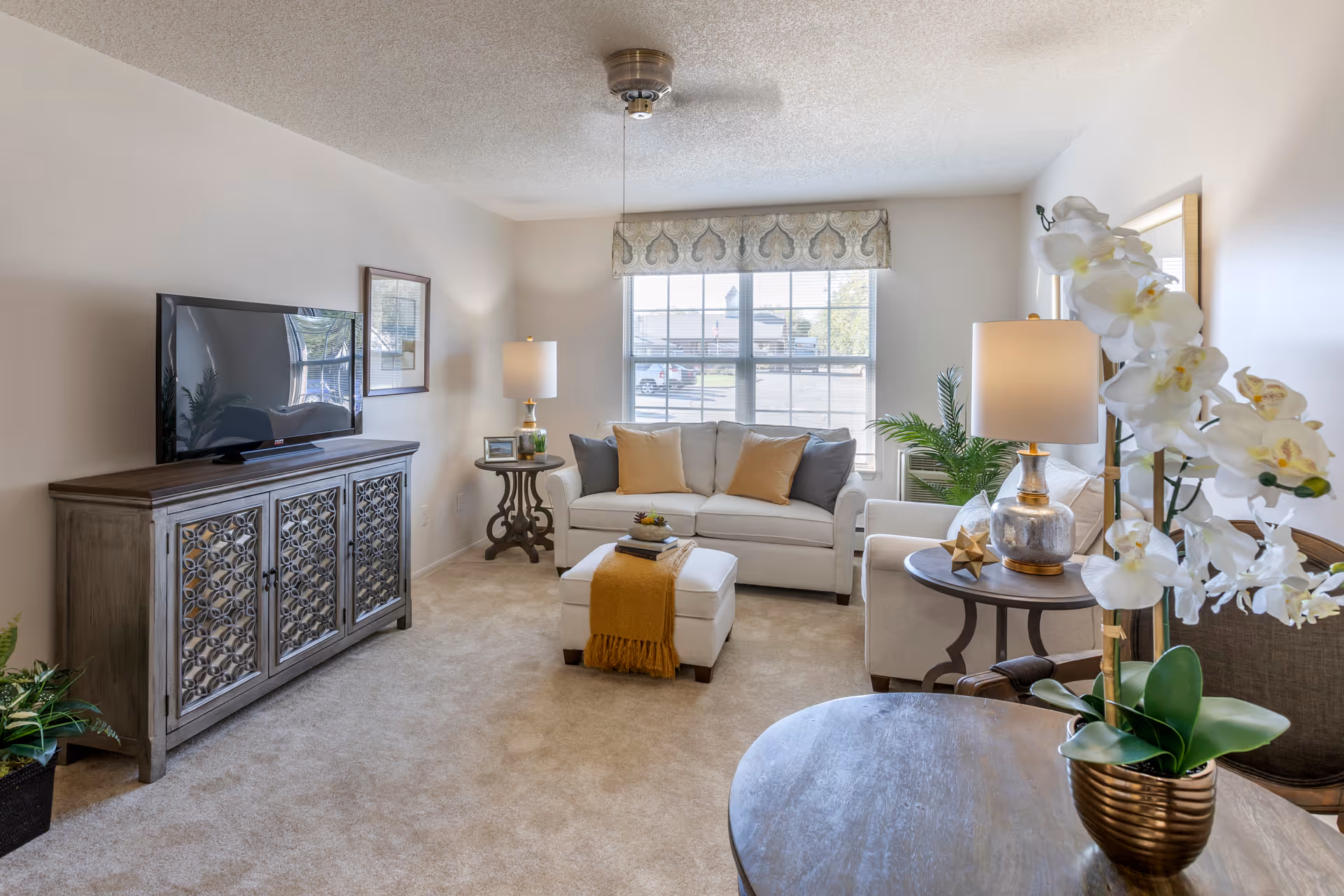 A bright and cozy living room with a beige carpet, a white sofa with yellow and gray cushions, a matching ottoman with a yellow throw, two side tables with lamps, a flat-screen TV on a decorative wooden cabinet, a large window with a patterned valance, and a round table with a white orchid plant in a gold pot.