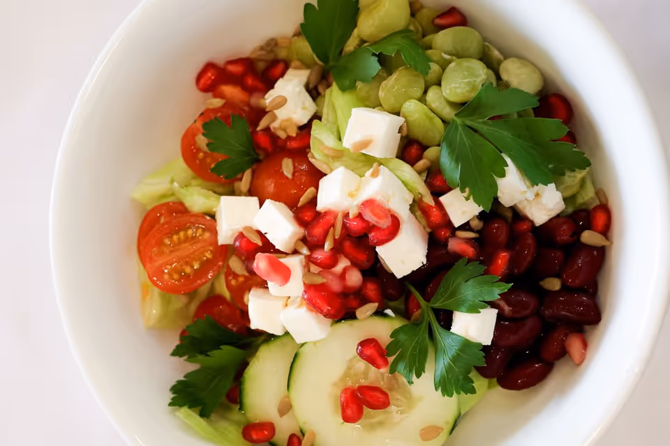 A white bowl filled with a fresh salad containing cherry tomatoes, cucumber slices, kidney beans, broad beans, feta cheese cubes, pomegranate seeds, sunflower seeds, and garnished with parsley leaves.