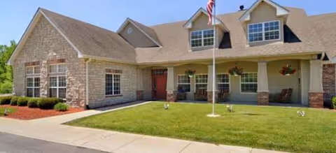 Front exterior of a senior living facility with a flagpole, porch seating, and manicured lawn.