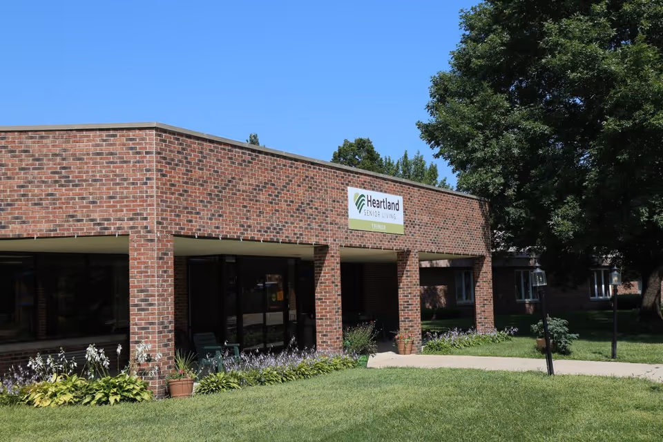 Exterior view of a single-story brick building with a sign that reads 'Heartland Senior Living Truman'. The building has large windows and a covered entrance area. There are plants and flowers along the building's edge and a large tree to the right. The sky is clear and blue.