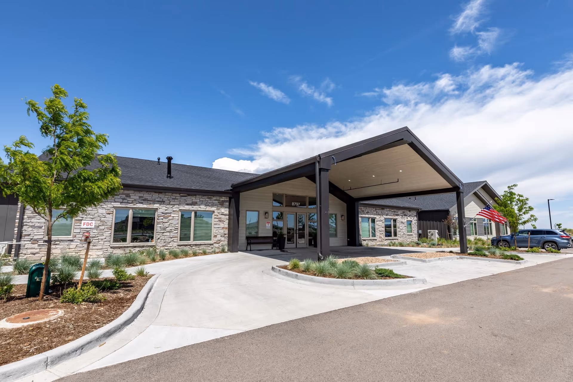 Exterior view of Terra Bluffs facility showing a modern single-story building with stone and siding facade, a covered entrance with a peaked roof, landscaped greenery, and an American flag near parked cars under a partly cloudy blue sky.