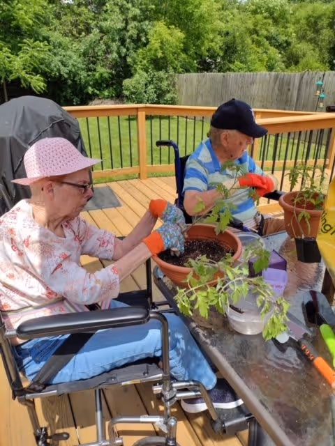 Two elderly individuals in wheelchairs are gardening on a wooden deck. They are wearing gloves and tending to potted plants on a glass table. One person is wearing a pink hat and floral shirt, while the other is wearing a blue striped shirt and a dark cap. The background shows green trees and a wooden fence.