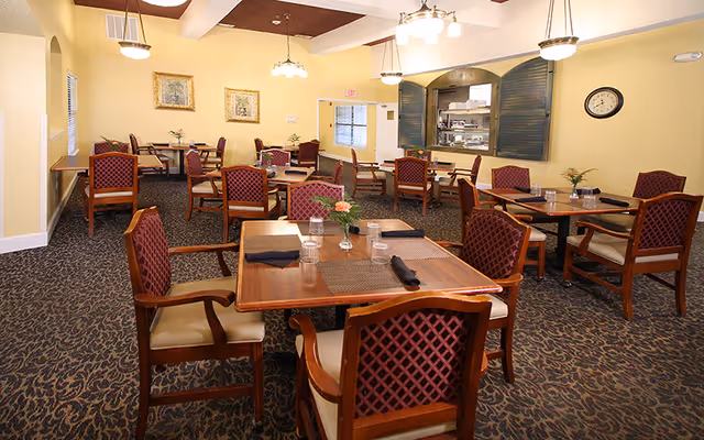 A dining room with multiple wooden tables and chairs arranged neatly. Each table has placemats, glasses, and napkins, with small flower arrangements as centerpieces. The room has yellow walls, carpeted flooring with a patterned design, and ceiling lights. There is a serving window with shutters on one wall and framed artwork hanging on another.