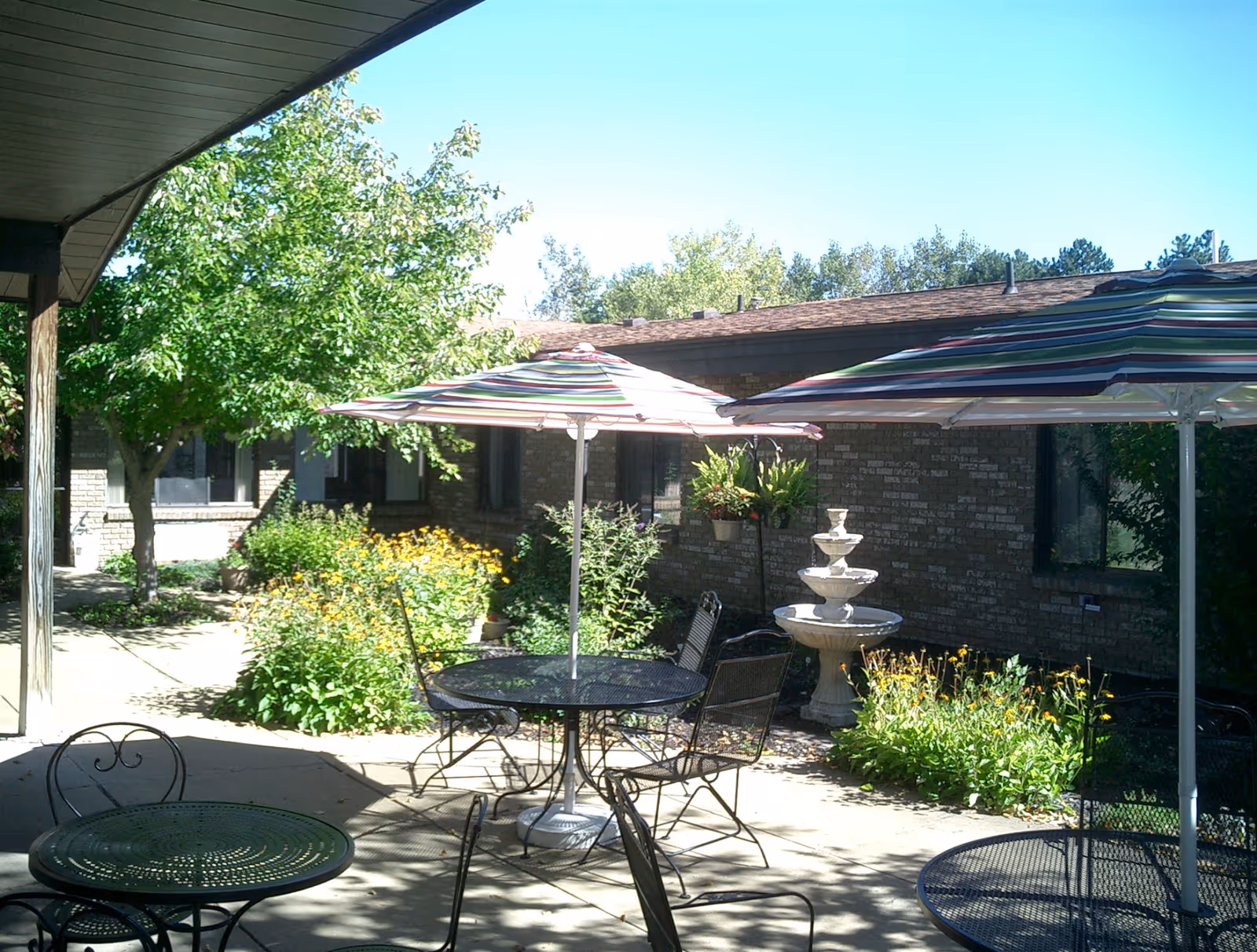 Outdoor patio area with metal tables and chairs, some tables shaded by striped umbrellas. There are green plants, yellow flowers, a tree, and a white tiered fountain near a brick building under a clear blue sky.