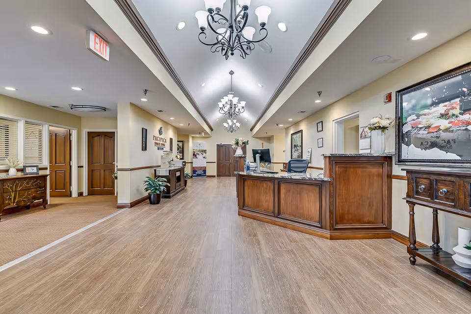 A spacious reception area with wooden flooring and a wooden reception desk on the right side. The ceiling features elegant chandeliers and recessed lighting. There are framed pictures and decorative plants placed around the room. Doors and a hallway are visible in the background, with an exit sign on the left side.