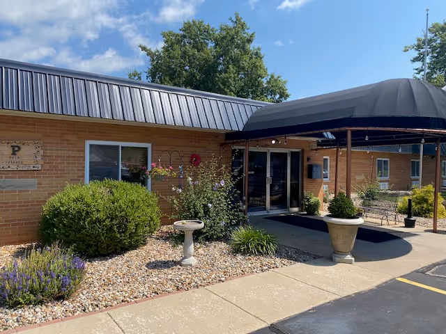 Exterior view of Palm Garden of Mattoon facility showing a brick building with a black metal roof and a covered entrance. There are bushes, flowers, and a birdbath in a landscaped area with rocks near the sidewalk leading to the entrance. The sky is partly cloudy.