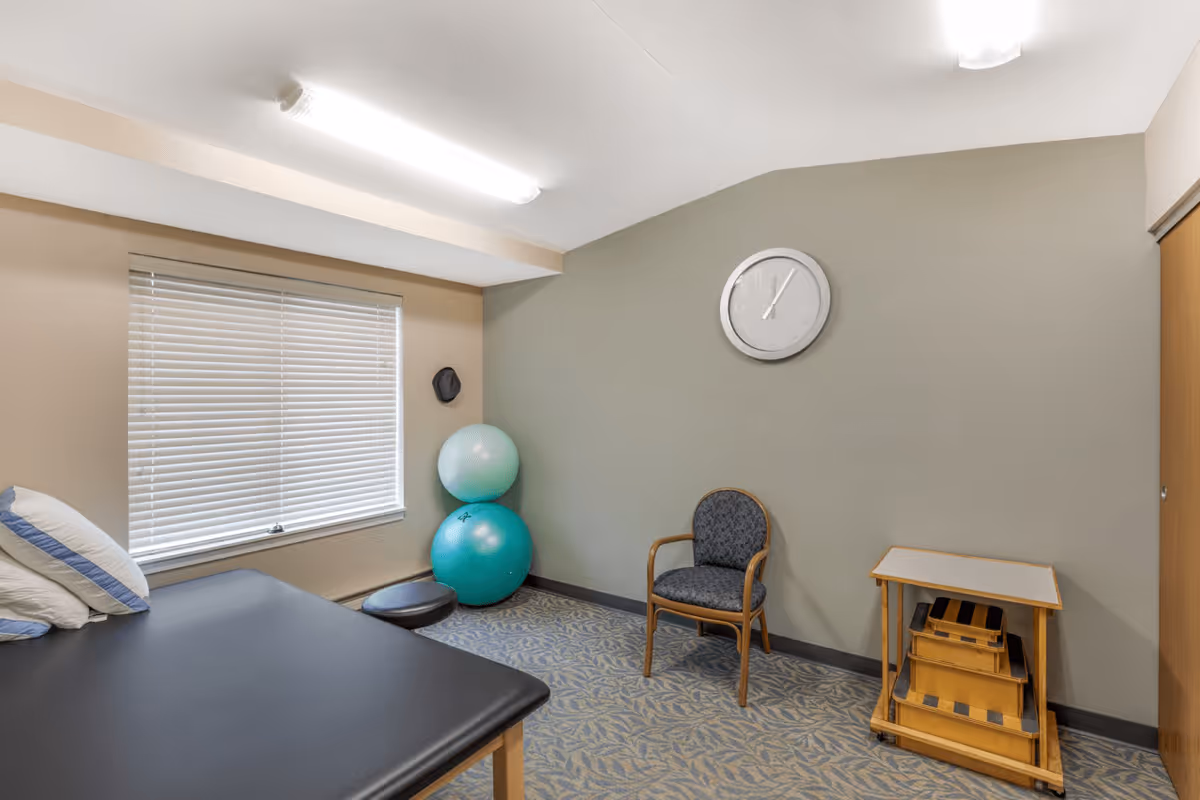 A small therapy or exercise room with a black padded table on the left, two large exercise balls stacked in the corner, a single chair against a light green wall, a small wooden step stool, a clock showing 12:05, and a window with closed blinds.