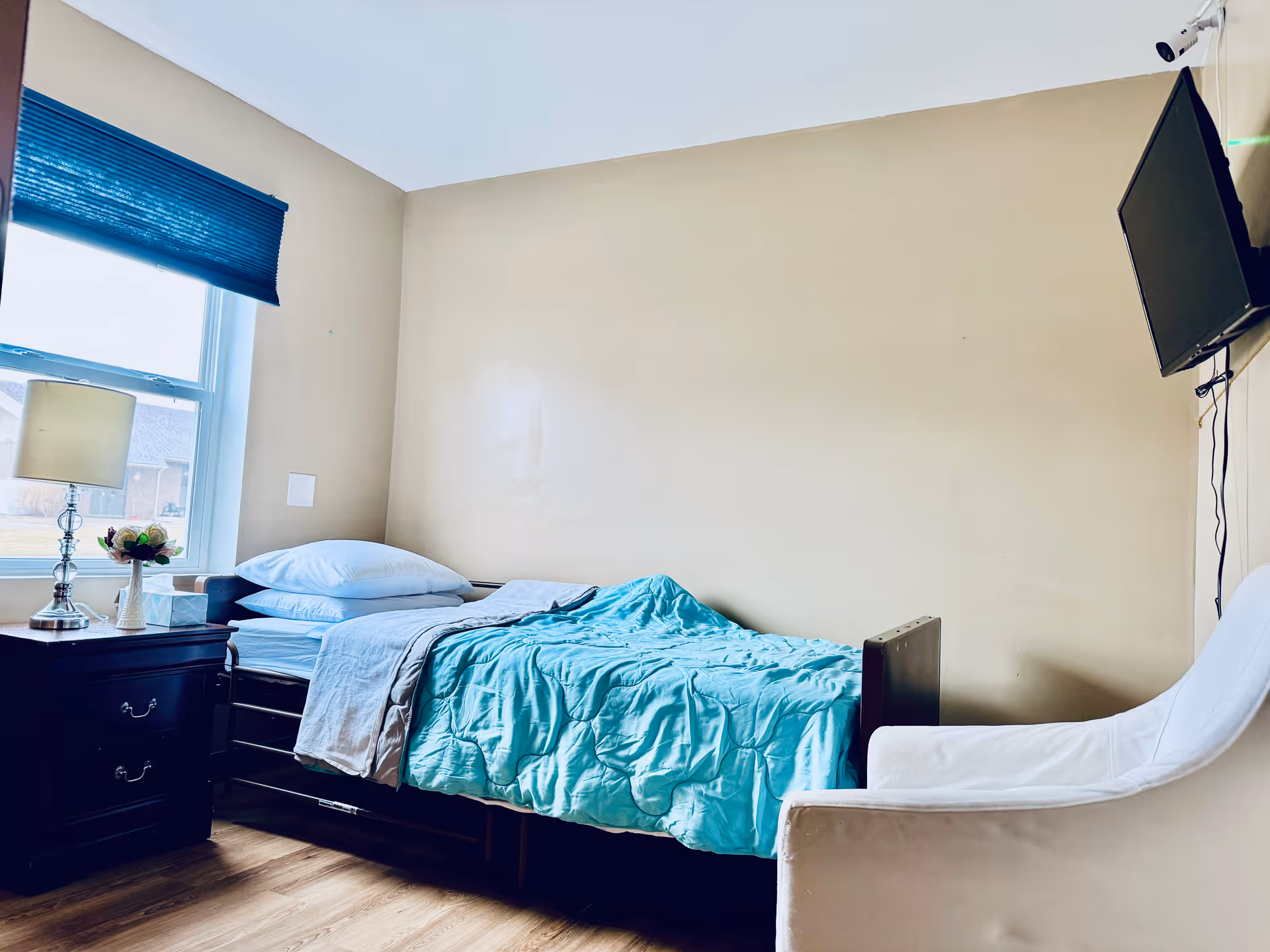 A small bedroom with a single bed covered with a light blue blanket and white pillows. Next to the bed is a dark wooden nightstand with a lamp, a small flower arrangement, and a tissue box. A window with a blue blind is on the left wall, and a white armchair is positioned near the foot of the bed. A flat-screen TV is mounted on the right wall.