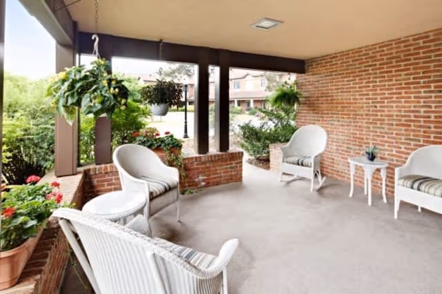 Covered outdoor patio area with white wicker chairs and a small white table. The patio has hanging plants and potted flowers, with a brick wall and columns. In the background, there are shrubs and a view of a building.