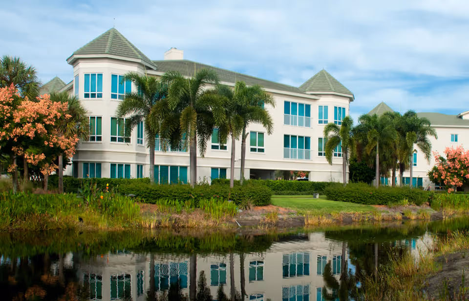 Exterior view of a multi-story senior living facility with green roofs and large windows, surrounded by palm trees, flowering trees, and greenery, reflected in a calm body of water in the foreground under a partly cloudy sky.