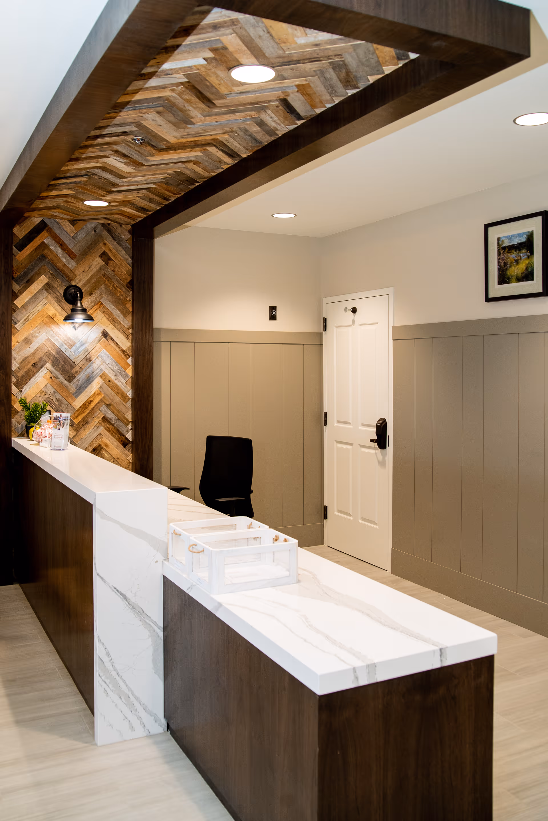 Reception area with a modern wooden and white marble desk, a black office chair behind the desk, wood-patterned ceiling and wall paneling, a white door, and a framed picture on the wall.