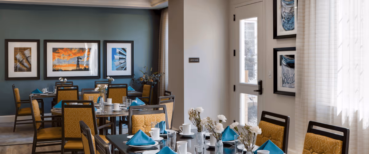 A dining room with multiple tables set for a meal, featuring yellow cushioned chairs, white cups, glasses, and blue folded napkins. The walls are decorated with framed artwork, and natural light enters through a window with sheer curtains on the right side.