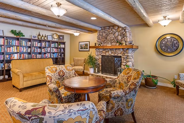 Cozy senior living facility common area with a stone fireplace, wooden ceiling beams, a round wooden table surrounded by four floral-patterned armchairs, a mustard yellow sofa, a bookshelf filled with books, potted plants, and a large wall clock.