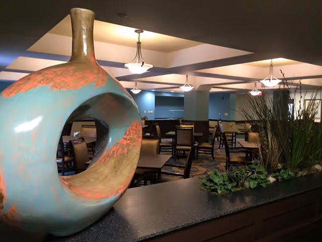 Dining room interior with tables and chairs, a large round ceramic sculpture on a counter and plants in the foreground.