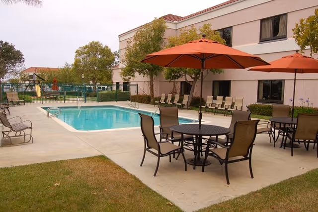 Outdoor area of a senior living facility featuring a swimming pool surrounded by a concrete deck with several lounge chairs and tables with orange umbrellas. The building with windows and some trees are visible in the background.